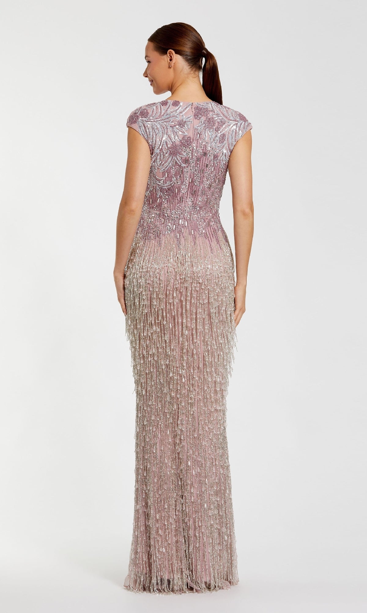 A woman stands facing away, wearing the Mac Duggal 6169 Beaded Long Lilac Formal Dress with intricate fringe and a floor-length skirt. Her hair is in a low ponytail, completing the elegant look.